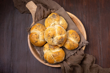 Top View of Homemade Kaiser Rolls Topped with Poppy Seeds in a Round Wooden Bowl on a Wooden Table