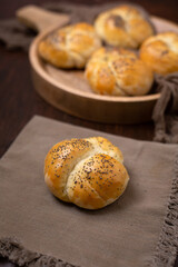 Homemade Kaiser Roll Topped with Poppy Seeds  on a Brown Napkin; More in a Round Wooden Bowl in the Background on a Wooden Table