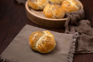 Homemade Kaiser Roll Topped with Poppy Seeds  on a Brown Napkin; More in a Round Wooden Bowl in the Background on a Wooden Table