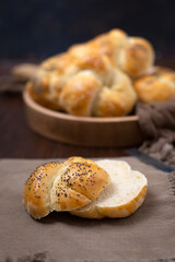 Close Up of a Homemade Kaiser Roll Topped with Poppy Seeds Cut Open  on a Brown Napkin; More in a Round Wooden Bowl in the Background on a Wooden Table
