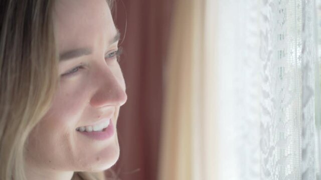 Hopeful caucasian woman smiles while looking out home window with white curtain
