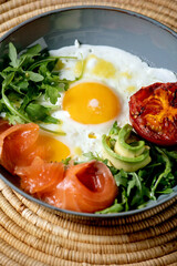 Healthy breakfast bowl with fried eggs, salmon, avocado, grilled tomato and salad serving with bread on straw napkin over dark marble background. Close up
