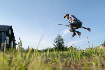 Funny young man in casual clothes flies on a broomstick, in the countryside, on a summer day. Positive lifestyle. Bottom shot.