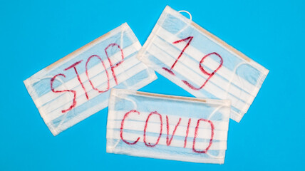 Typical 3-ply white surgical mask with rubber earhooks to cover mouth and nose with English block letters Stop Covid-19 on a blue background. Dangerous virus and quarantine concept