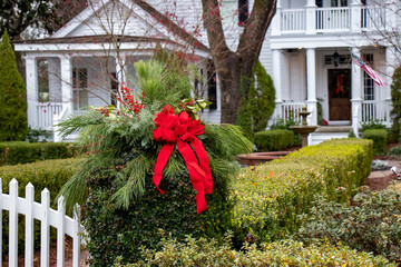 Close-up of natural Christmas wreath of evergreens, berries, and red ribbon on an ivy-covered gate post at the entrance to the front garden of a beautiful white house.