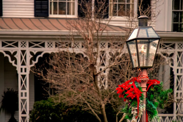 Close-up of an old gas streetlamp decorated for the Christmas holiday with ribbons and evergreens burning brightly at twilight in front of a Victorian style house.