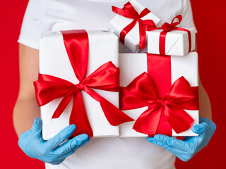 Holiday celebration. Gift surprise. Covid-19 protection. Unrecognizable woman in medicine gloves holding present boxes wrapped taped ruby ribbon isolated on red. Happy birthday. Family greetings.