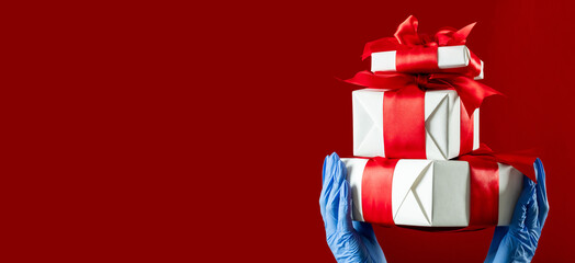 Christmas gifts. Covid-19 protection. Greeting boxes. Safety delivery. Holidays 2021. Hands in medicine gloves holding wrapped presents taped ruby ribbon isolated on red copy space.