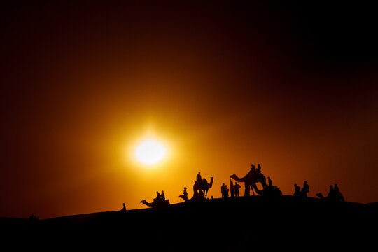 Silhouettes of camel caravan with people walking in dark desert against sunset sky with bright sun shining over horizon in India