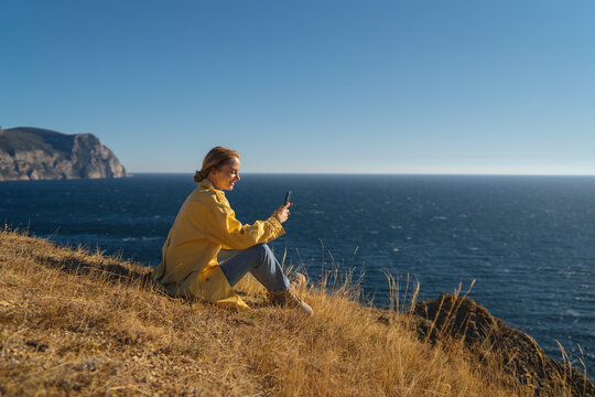 A girl sits on a high seashore and makes a frame on the phone