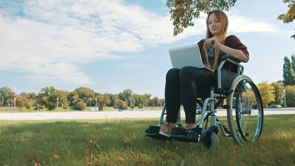 Remote work concept. Young beautiful disabled caucasian woman in wheelchair using laptop in nature. High quality photo