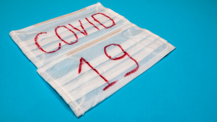Typical white 3-ply surgical mask with rubber earhooks to cover mouth and nose with English block letters Covid-19 on a blue background. Dangerous virus and quarantine concept.