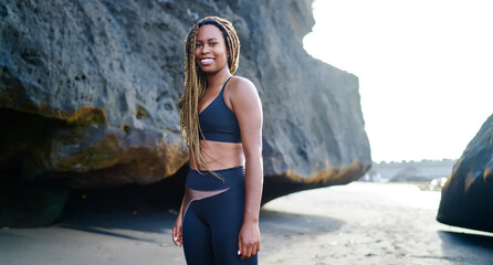 Smiling ethnic woman in sportswear standing along on seashore and looking at camera
