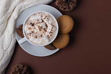 Autumn and winter concept. Top view of cup of cocoa with marshmallows, cookies, white sweater and pine cones on brown background.