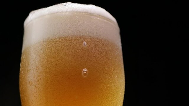 Low angle of soft white foam rising and spilling from glass cup of cold beer on black background