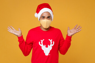 Fototapeta na wymiar Confused young Santa african american man in Christmas hat face mask to safe from coronavirus virus covid-19 spreading hands isolated on yellow background studio. New Year celebration holiday concept.