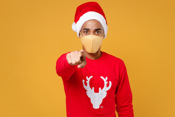 Fototapeta na wymiar Santa african american man in Christmas hat face mask to safe from coronavirus virus covid-19 pointing index finger on camera isolated on yellow background. Happy New Year celebration holiday concept.