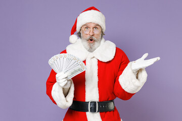 Fototapeta na wymiar Shocked Santa Claus man in Christmas hat red suit coat glasses hold fan of cash money dollar banknotes spreading hands isolated on violet background. Happy New Year celebration merry holiday concept.
