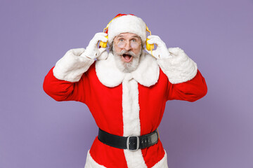 Fototapeta na wymiar Shocked Santa Claus man in Christmas hat red suit coat white gloves glasses listening music with headphones isolated on violet background studio. Happy New Year celebration merry holiday concept.