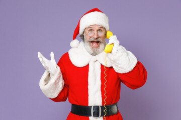 Surprised Santa Claus man in Christmas hat red suit coat gloves glasses talking on telephone spreading hands isolated on violet background studio. Happy New Year celebration merry holiday concept.