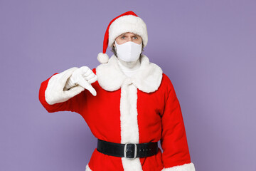 Santa Claus man in Christmas hat suit mask to safe from coronavirus virus covid-19 during quarantine showing thumb down isolated on violet background. Happy New Year celebration merry holiday concept.