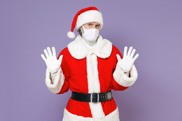 Fototapeta na wymiar Santa Claus man in Christmas hat red suit face mask to safe from coronavirus virus covid-19 rising hands up showing palms isolated on violet background. New Year celebration merry holiday concept.