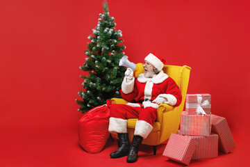 Crazy Santa Claus man in Christmas hat suit glasses sit in armchair with fir tree presents gifts screaming in megaphone isolated on red background. Happy New Year celebration merry holiday concept.