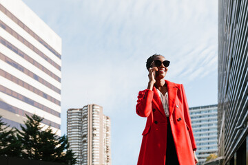 Enthusiastic black woman in red coat discussing on smartphone in downtown