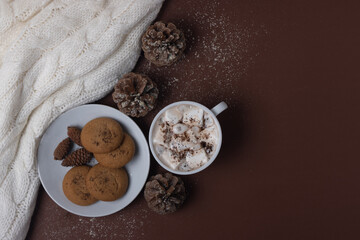 Top view of cup of cocoa with marshmallows, cookies, white sweater, pine cones and snow on brown background. Autumn and winter concept. Chestnut color.