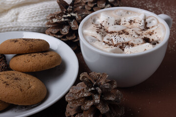 Cup of cocoa with marshmallows, cookies, white sweater, pine cones and snow on brown background. Autumn and winter concept.  Chestnut color.