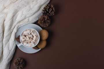 Autumn and winter concept. Top view of cup of cocoa with marshmallows, cookies, white sweater and pine cones on brown background. Chestnut color