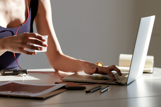 Faceless woman drinking water while working with papers and using laptop