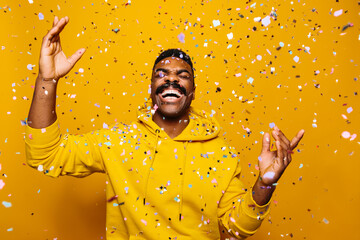Happy african american man over yellow background with confetti falling on head