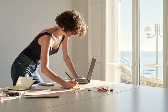 Calm slim woman taking notes in notepad while working on project at home
