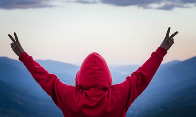 Unrecognizable woman from behind with red sweatshirt and hood raises her arms in front of the mountains. Adventurous and independent woman