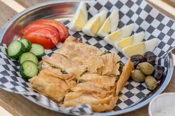 Top view of a plate, with homemade traditional turkish style Tabla Borek, boiled eggs, yogurt, sliced vegetables and olives, over a black and white paper napkin.