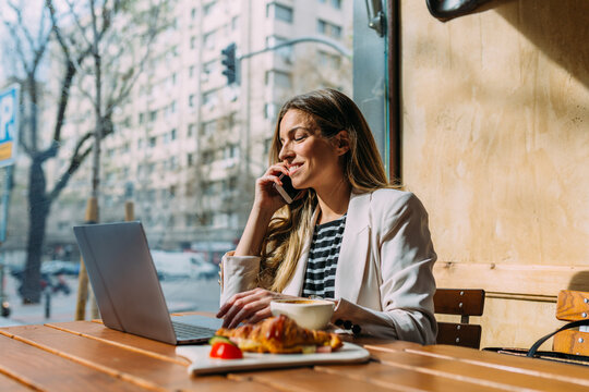 Confident businesswoman using cellphone and laptop in cafeteria.
