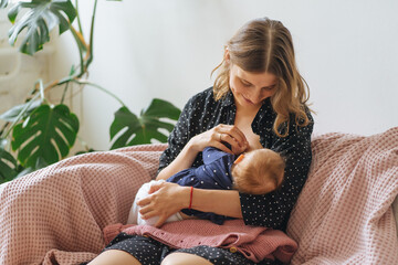 young beautiful mother breastfeeds her baby sitting on a sofa