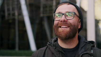 Close up view of cheerful guy in glasses having good mood and smiling while walking at city street. Portrait of bearded man looking happy and laughing. Concept of emotions.