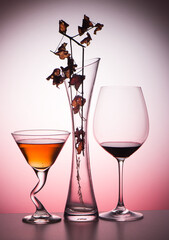 Autumn still life with a glass of red wine and a glass of cider. Vase with a branch with dry leaves. Light colored background.