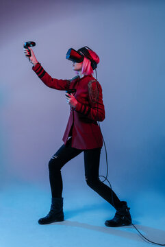 A girl with pink hair in a red jacket and VR Headset in the color light plays with controllers some video games