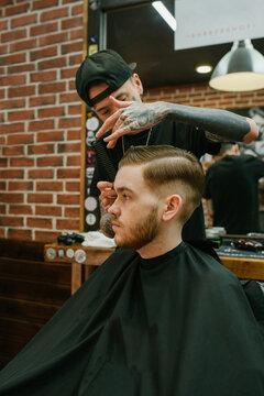 Ginger haired guy sits on the chair in the barbershop and tattoed barber works with his hair