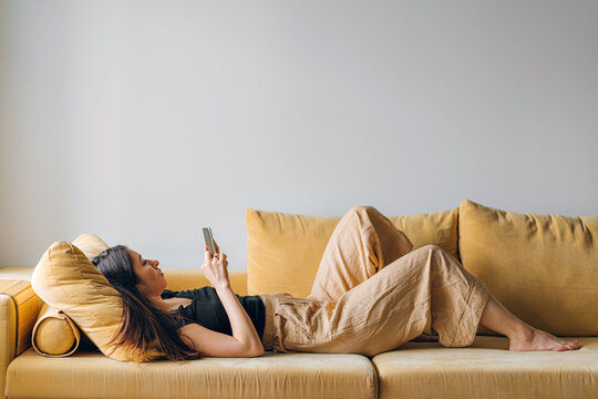 side view of a young brunette lying with a smartphone in her hands on a yellow sofa