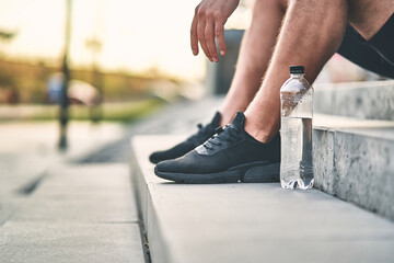 In focus a transparent plastic bottle with water stands on the stairs near the male feet in black sneakers. Sport concept