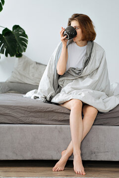 young woman is drinking coffee while sitting on the edge of her bed