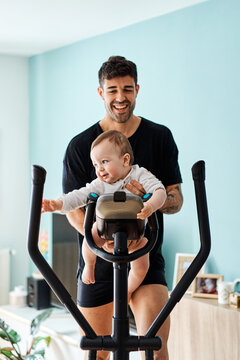Young father doing sport at home with his baby in an elliptical bike