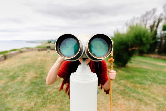 Unrecognizable female hiker with trekking stick looking through binocular telescope while standing on green meadow near sea and exploring nature during pilgrimage through Spain