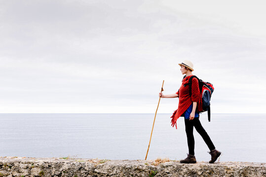 Full body side view of female hiker with backpack and trekking stick standing on stone cliff border against gray cloudy sky and enjoying freedom during pilgrimage through Spain