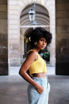 Side view of serious millennial African American female with curly hair and earrings wearing yellow crop top and blue jeans looking at camera while standing against old stone building on city street