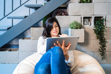 Focused young female designer in casual clothes sitting on comfortable pouf and using graphic tablet while working in contemporary creative workspace